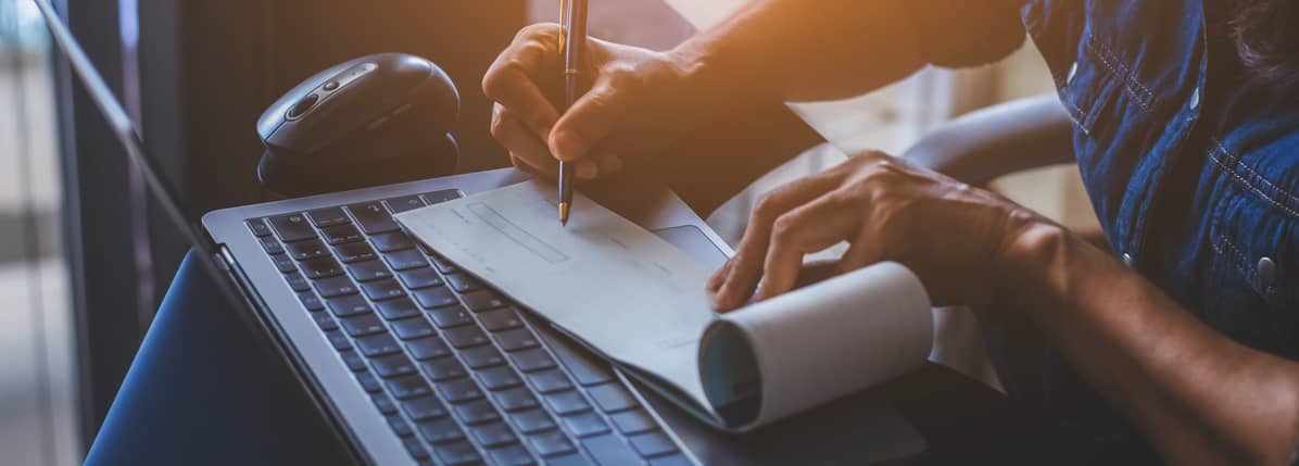 Person writing a check on a notepad while using a laptop